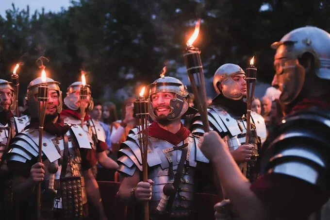 Group of people dressed as Roman soldiers holding torches during a historical reenactment or parade at dusk.