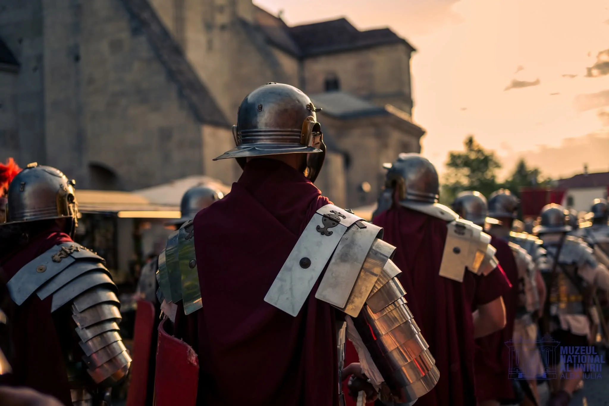 Group of people dressed as Roman soldiers with metallic armor and helmets, standing outdoors at sunset.
