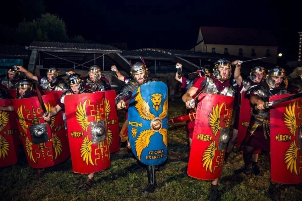Reenactors dressed as Roman soldiers with shields and helmets, participating in a reenactment event at night.