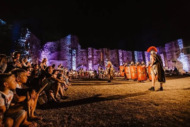A nighttime outdoor theatrical performance with actors in costume and an audience watching against a backdrop of illuminated stone walls or cliffs.