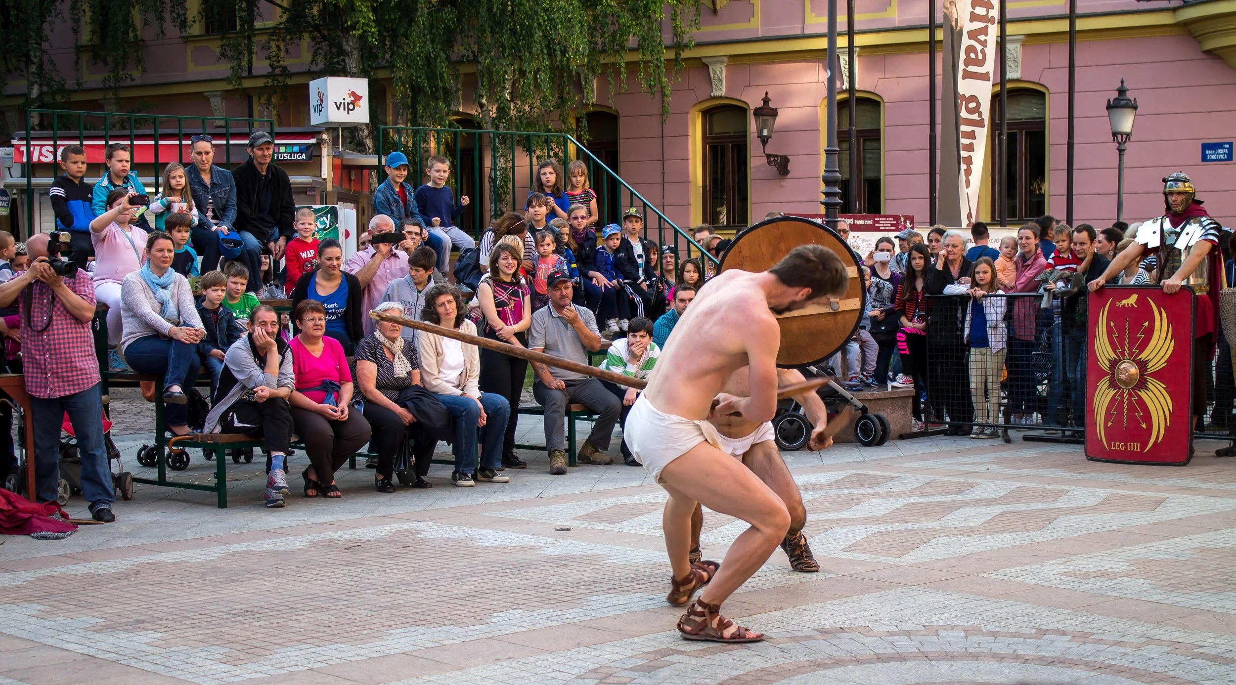 Two men dressed as ancient Roman gladiators performing a reenactment in front of a large crowd of spectators in an outdoor city square.