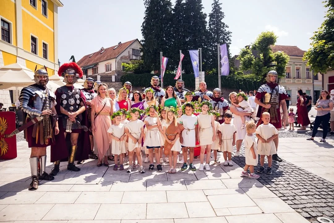Group of children and adults dressed in ancient Roman costumes, standing in a town square with historical buildings and flags in the background.