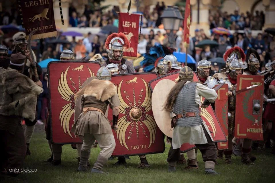 Historical reenactment of Roman soldiers in armor with shields, some with banners, on a grassy field during a festival or parade with a crowd of spectators in the background.