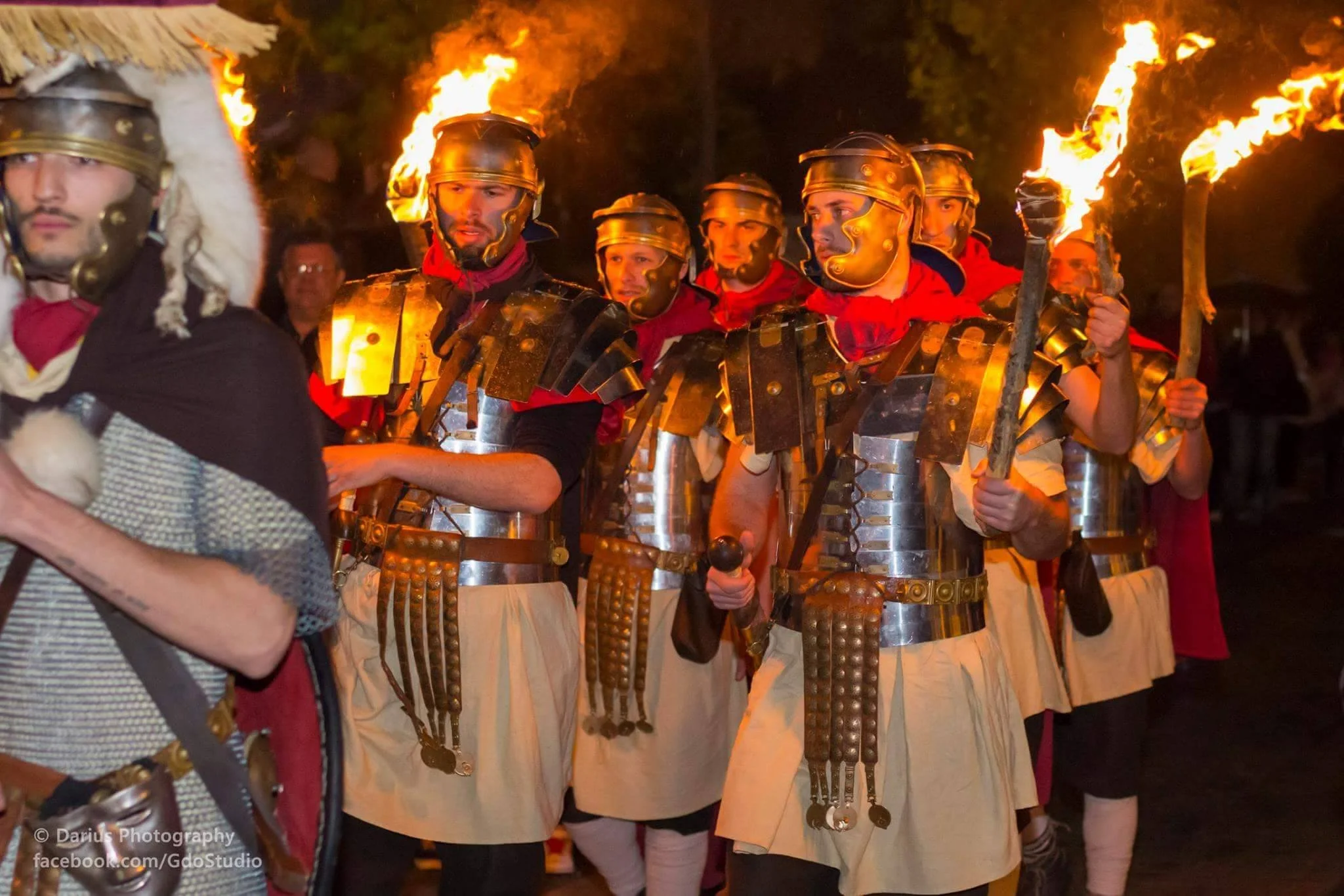A group of people dressed in Roman centurion costumes, carrying torches, participating in a nighttime parade or reenactment.