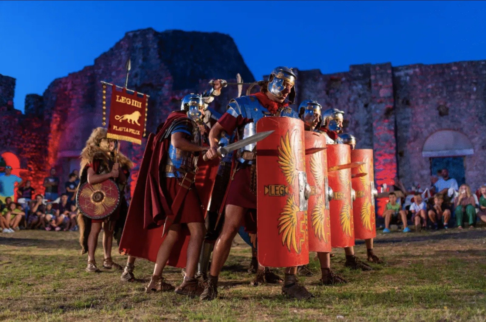 A group of people dressed as Roman soldiers participating in a reenactment or theatrical performance, with some holding shields and swords, and an audience in the background.