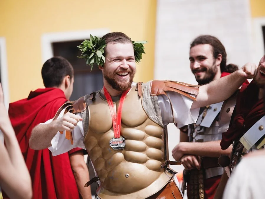 A man smiling, wearing a laurel wreath and a muscular costume, at a festive event with others in costumes.