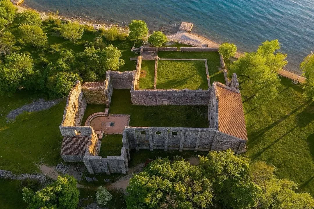 Aerial view of a historic stone castle ruins located near a body of water, surrounded by green trees and grass, with a small courtyard and a tiled roof section.