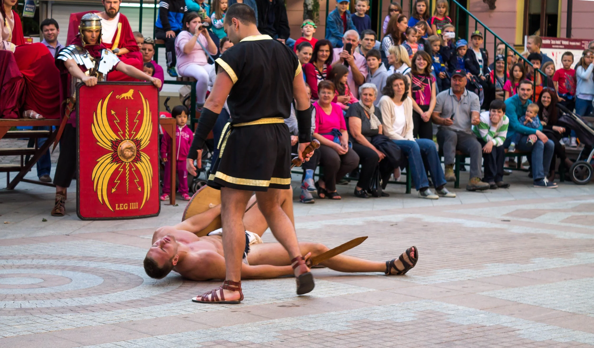 Street theater performance featuring two actors in Roman costumes, one lying on the ground and the other holding a sword, with a seated audience watching in the background.