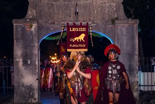 People dressed as Roman soldiers in a historical reenactment, with one holding a banner reading 'LEG IIII Felix'. The scene takes place under a stone archway, with crowd and torches visible in the background.