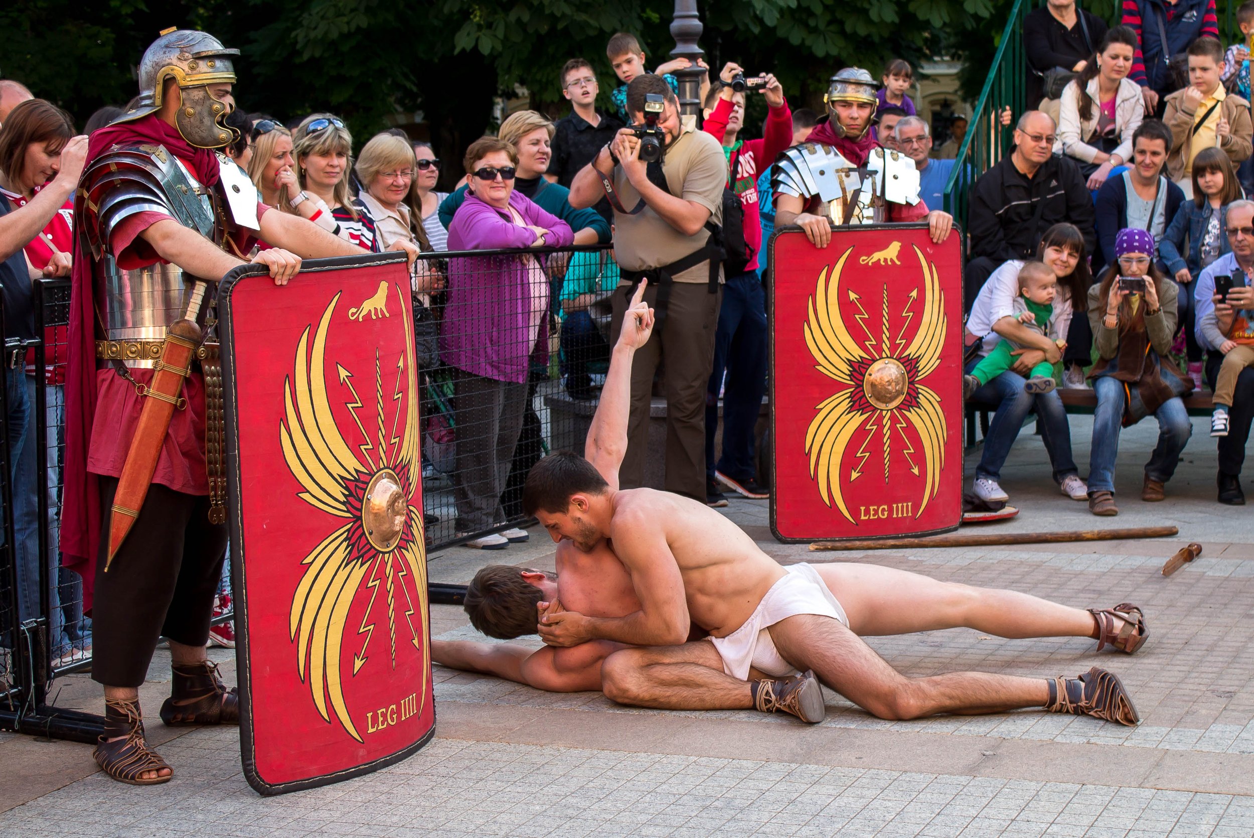 Two shirtless men wrestling on the ground in front of a crowd, with two men dressed as Roman soldiers holding shields, and people watching and filming at an outdoor event.