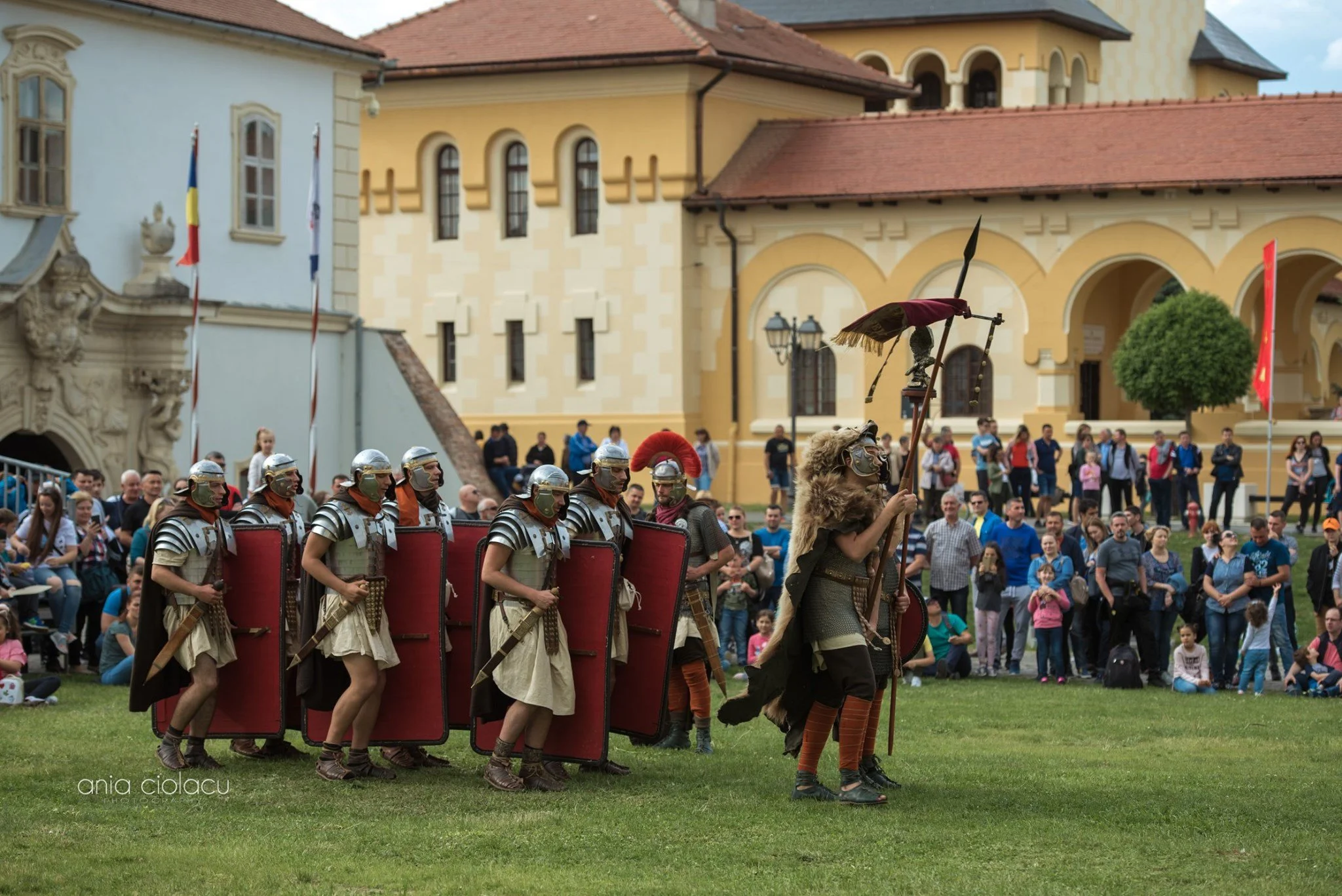 Group of people dressed as Roman soldiers and gladiators participating in a historical reenactment in front of a historic building with a crowd of spectators watching.