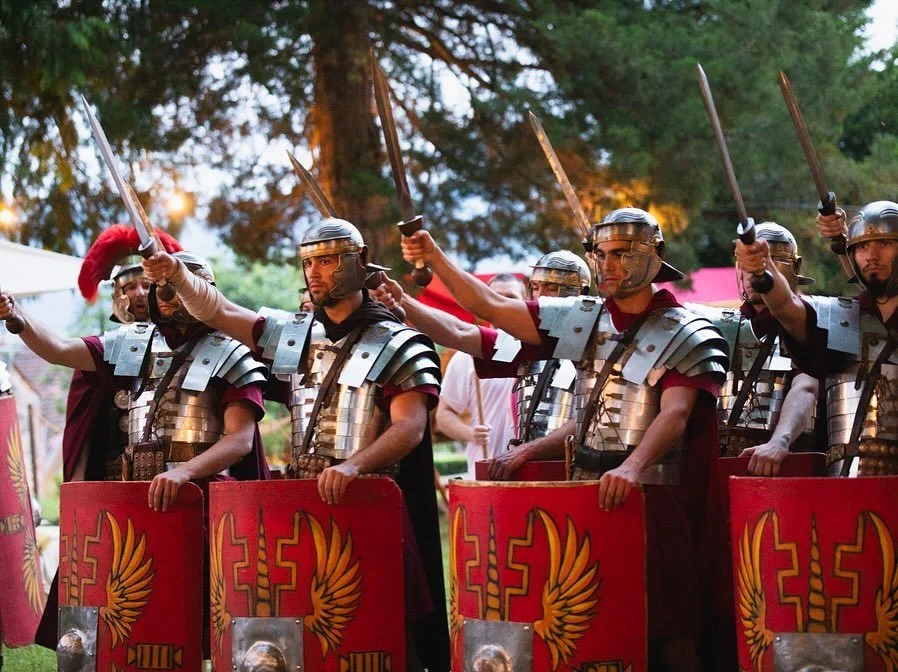 Group of men dressed as Roman soldiers holding shields and swords, standing outdoors with trees in the background.