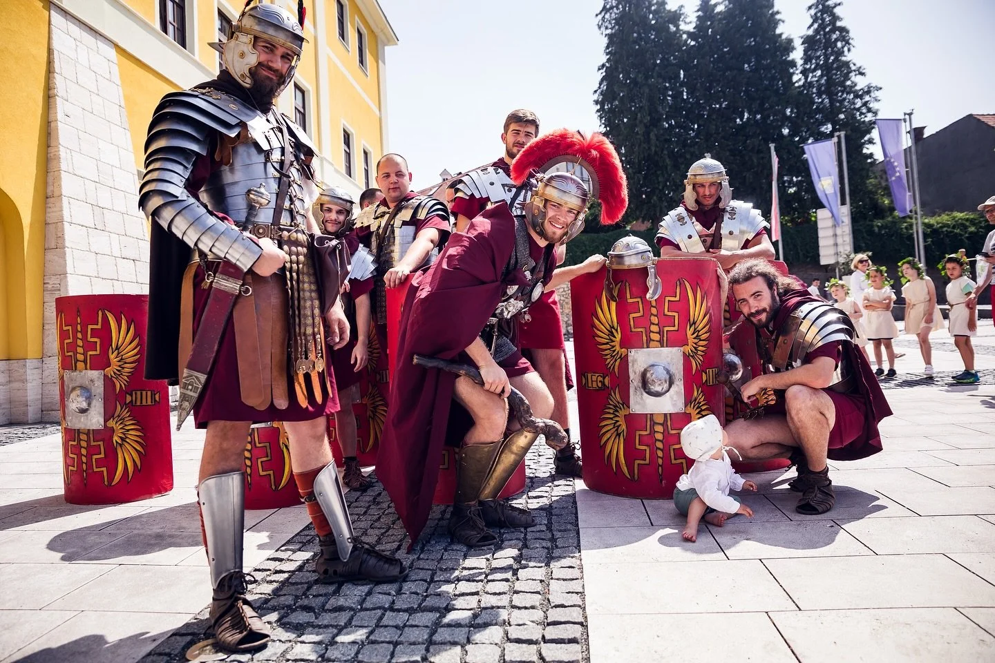 Group of people dressed as ancient Roman soldiers posing outdoors, with children in the background, in a sunny setting.