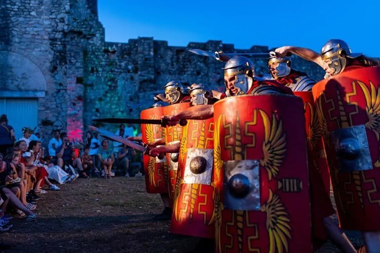 Group of people dressed as ancient Roman soldiers with shields and swords performing a reenactment for an audience outside during evening.