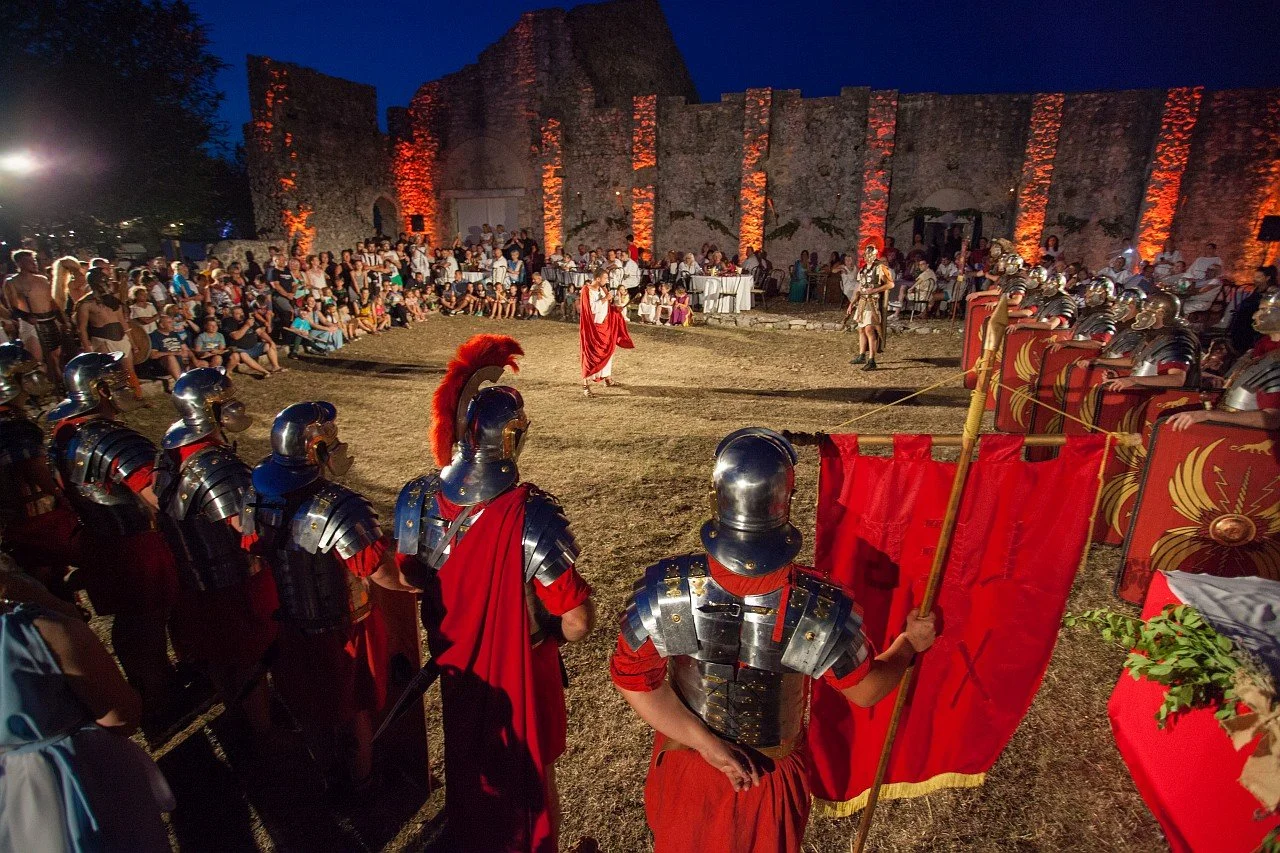 Roman reenactment at night featuring actors in armor, spears, and red cloaks, with an audience watching the performance outdoors near ancient stone ruins illuminated with orange lights.