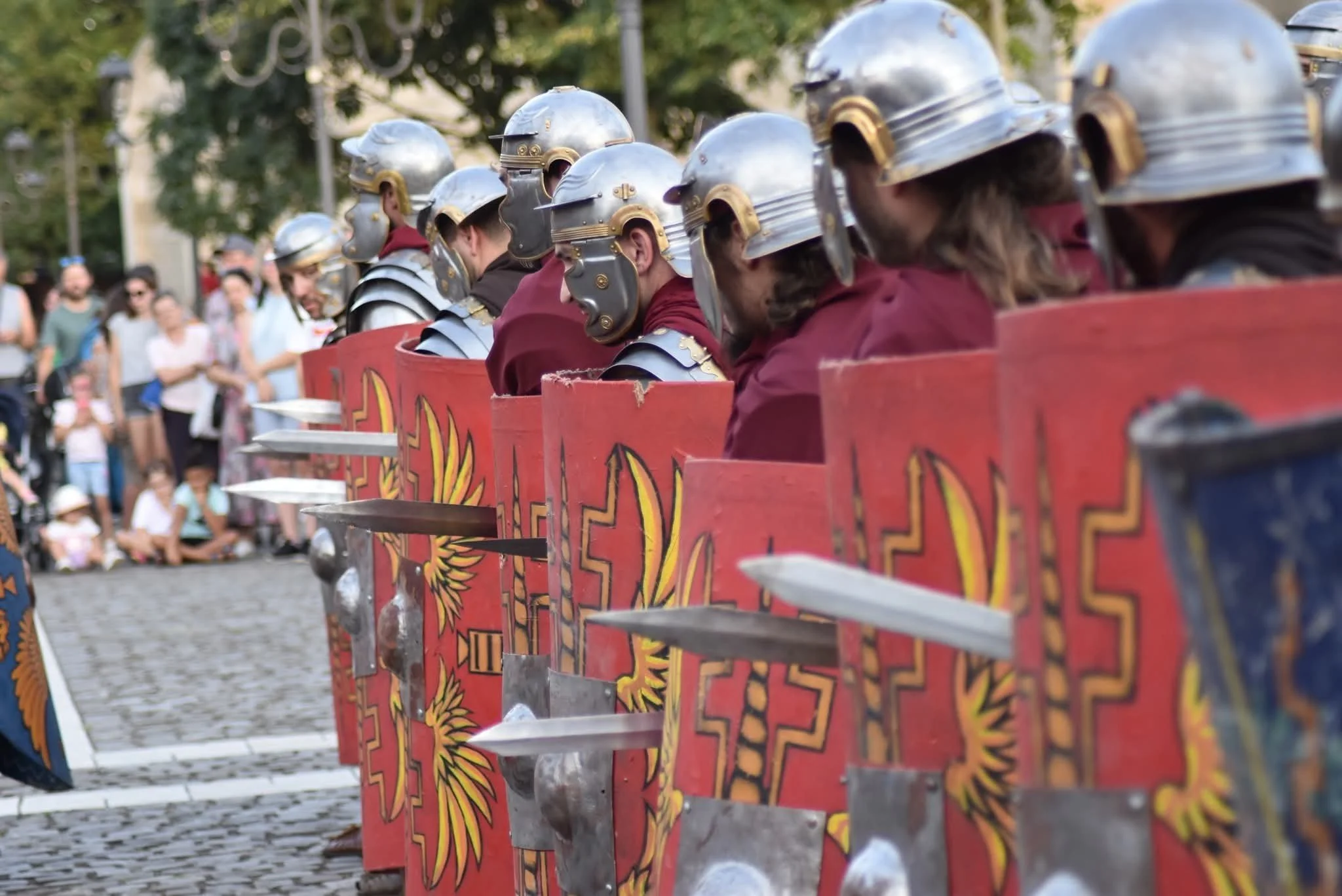 Line of people dressed as medieval soldiers in armor, holding red shields and swords, participating in a historical reenactment or parade.