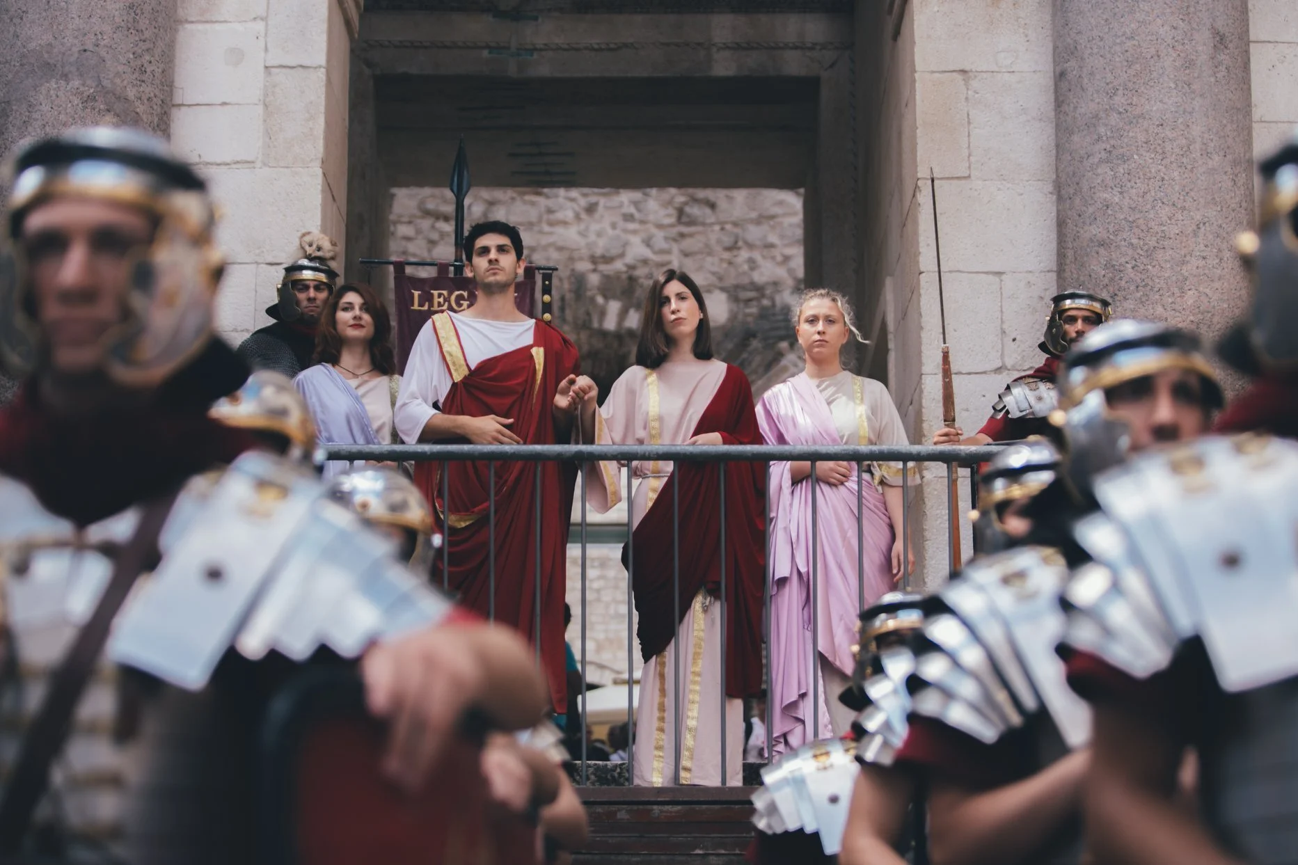 Historical reenactment with people dressed in ancient Roman-style clothing, standing on a balcony with soldiers in armor and helmets in the foreground.