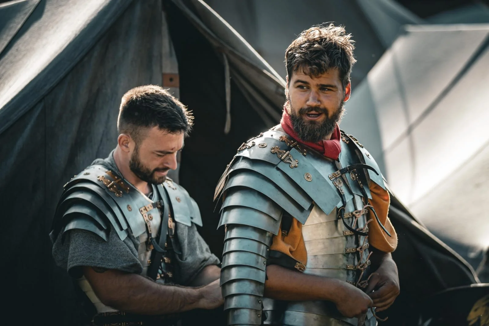 Two men dressed as Roman soldiers, wearing metallic armor, standing outdoors near a tent.