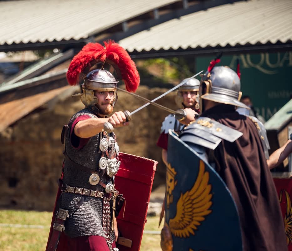 Two men dressed as Roman soldiers engaged in combat, wearing metal helmets with red plumes, armor, and holding shields and swords during a reenactment.