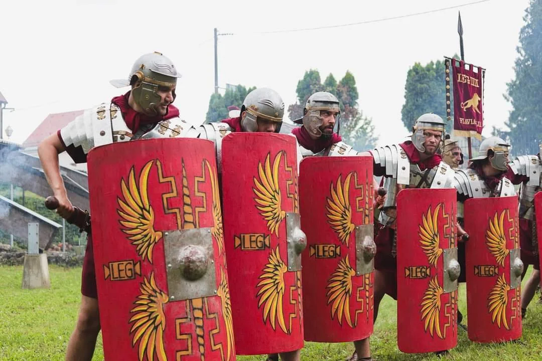 Group of people dressed as ancient Roman soldiers holding large red shields with yellow winged emblem and the word "LEG" on them, outdoors.