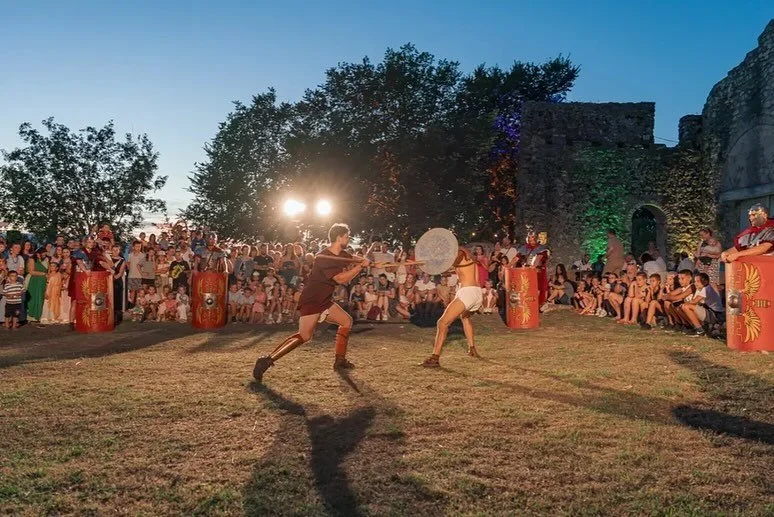 Two performers engaged in a medieval combat demonstration with shields and wooden swords in front of a large crowd at an outdoor event during evening hours.