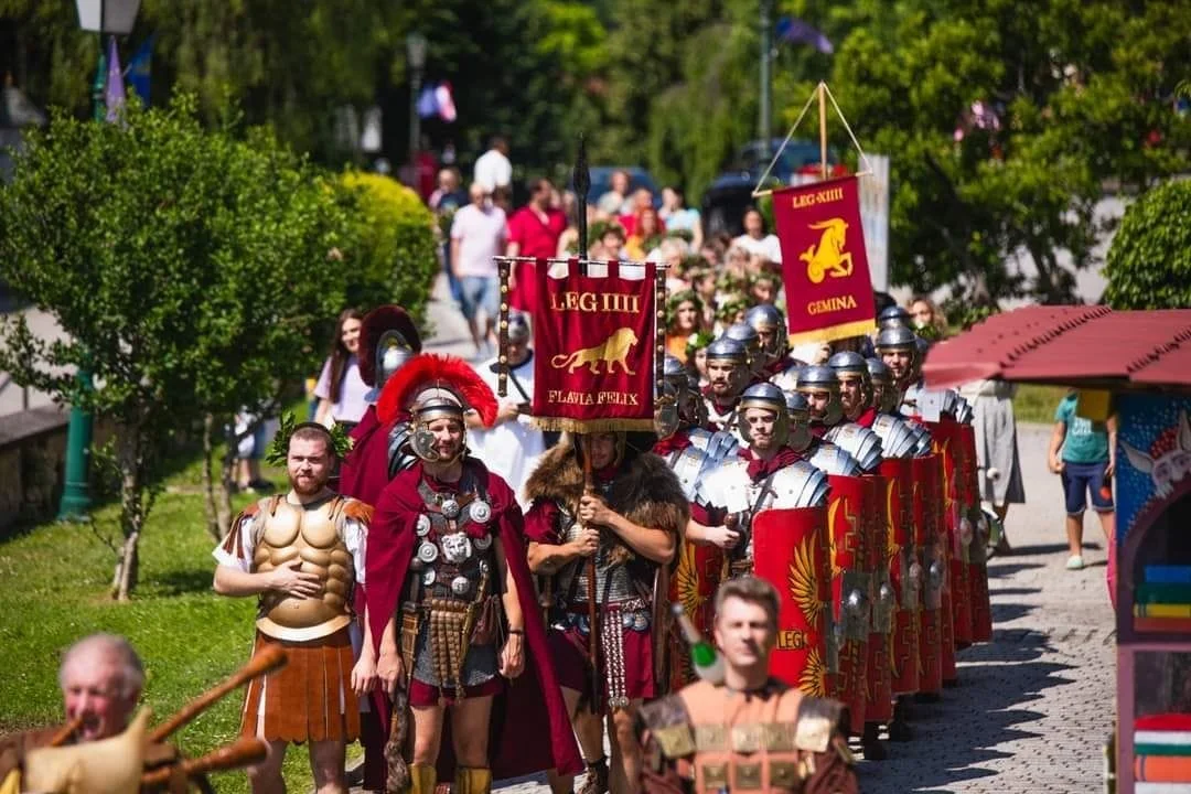 A reenactment parade featuring participants dressed as Roman soldiers, with some in armor carrying shields and banners, and others in tunics and helmets, walking along a pathway lined with trees and spectators.