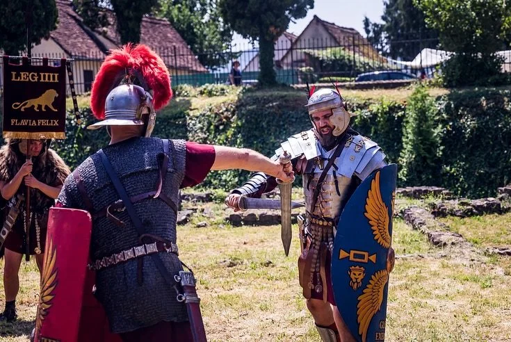 Two men dressed as Roman gladiators shaking hands during a reenactment or event outdoors, with a sign reading 'LEG IIII Flavia Felix' in the background.