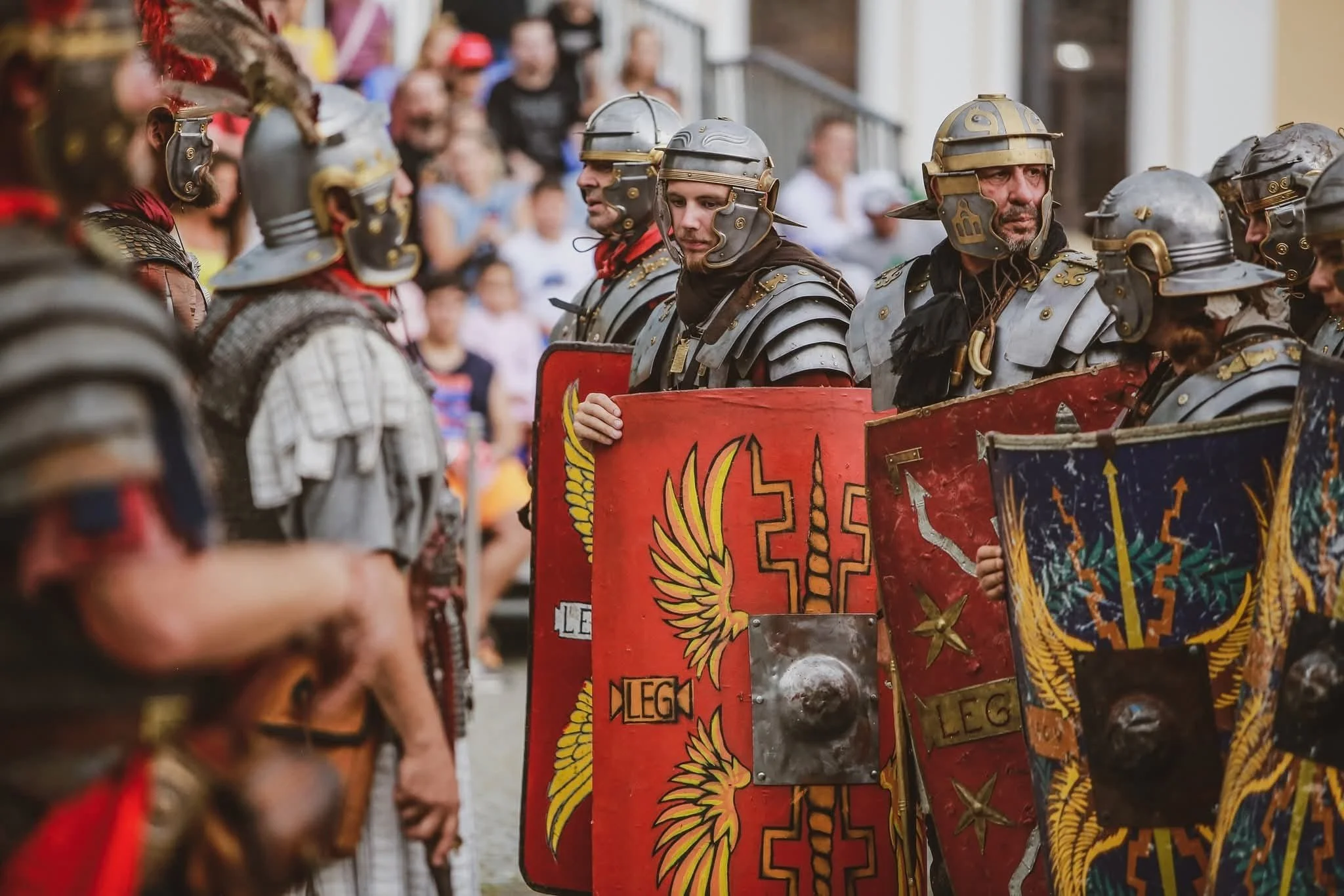Reenactors dressed as Roman soldiers holding shields during a historical parade, with a crowd of spectators in the background.