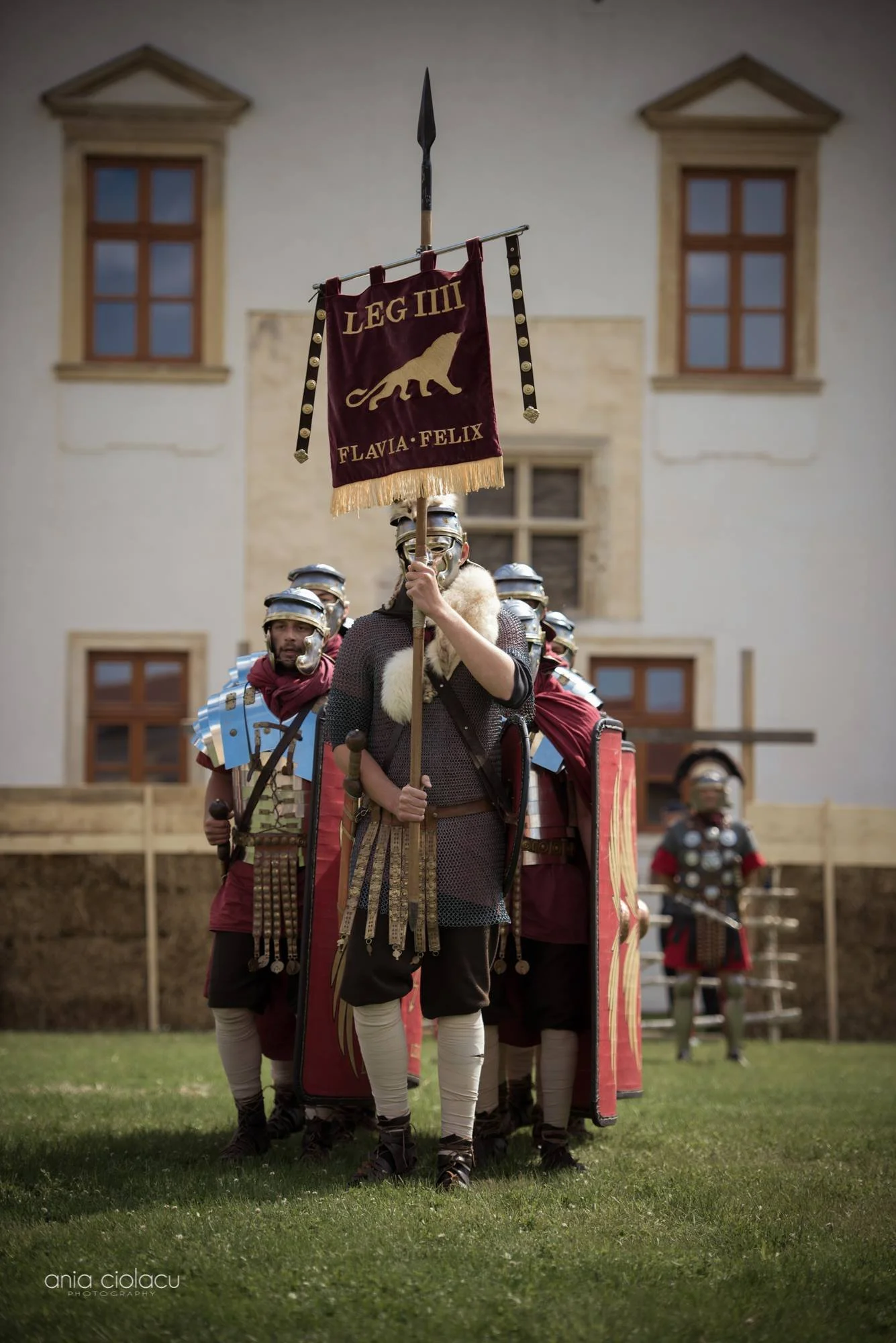 Group of people dressed as Roman soldiers in armor, with one person holding a banner with a lion and the words 'LEG IIII' and 'FLAVIA FELIX'. They are outdoors in a grassy area with a building in the background.