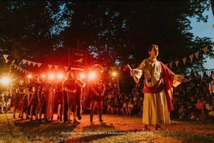 Young boy dressed as a king or historical figure standing on a stage at an outdoor event during dusk, with people sitting in the audience and others standing around. Bright stage lights illuminate the scene, and decorative bunting hangs overhead.
