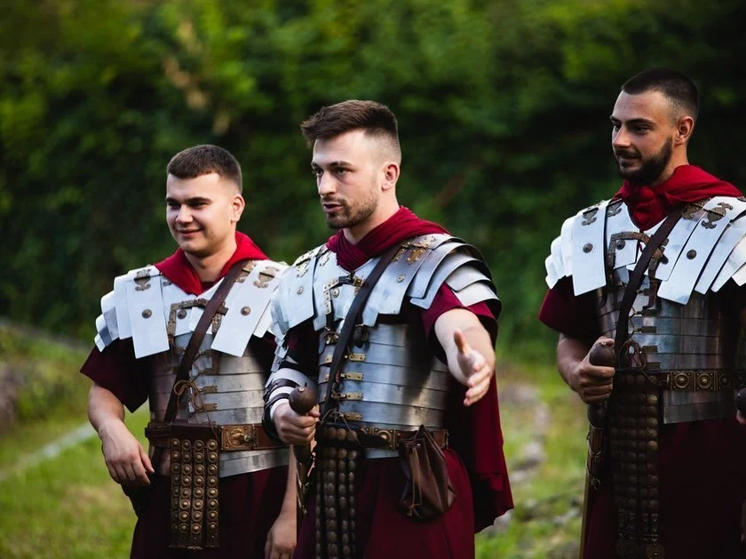 Three men dressed as Roman soldiers standing outdoors with green foliage in the background.