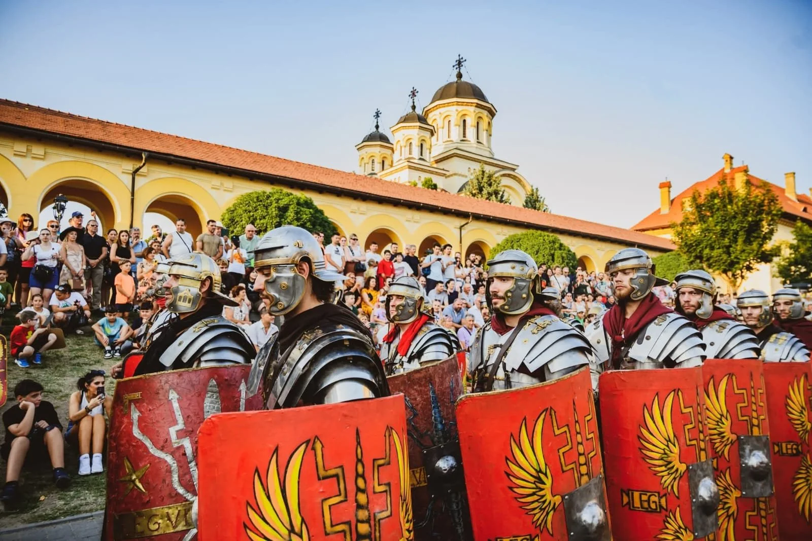 Roman soldiers in armor and shields parade in front of a crowd of spectators during a festival or reenactment, with historic buildings and a church with domes in the background.
