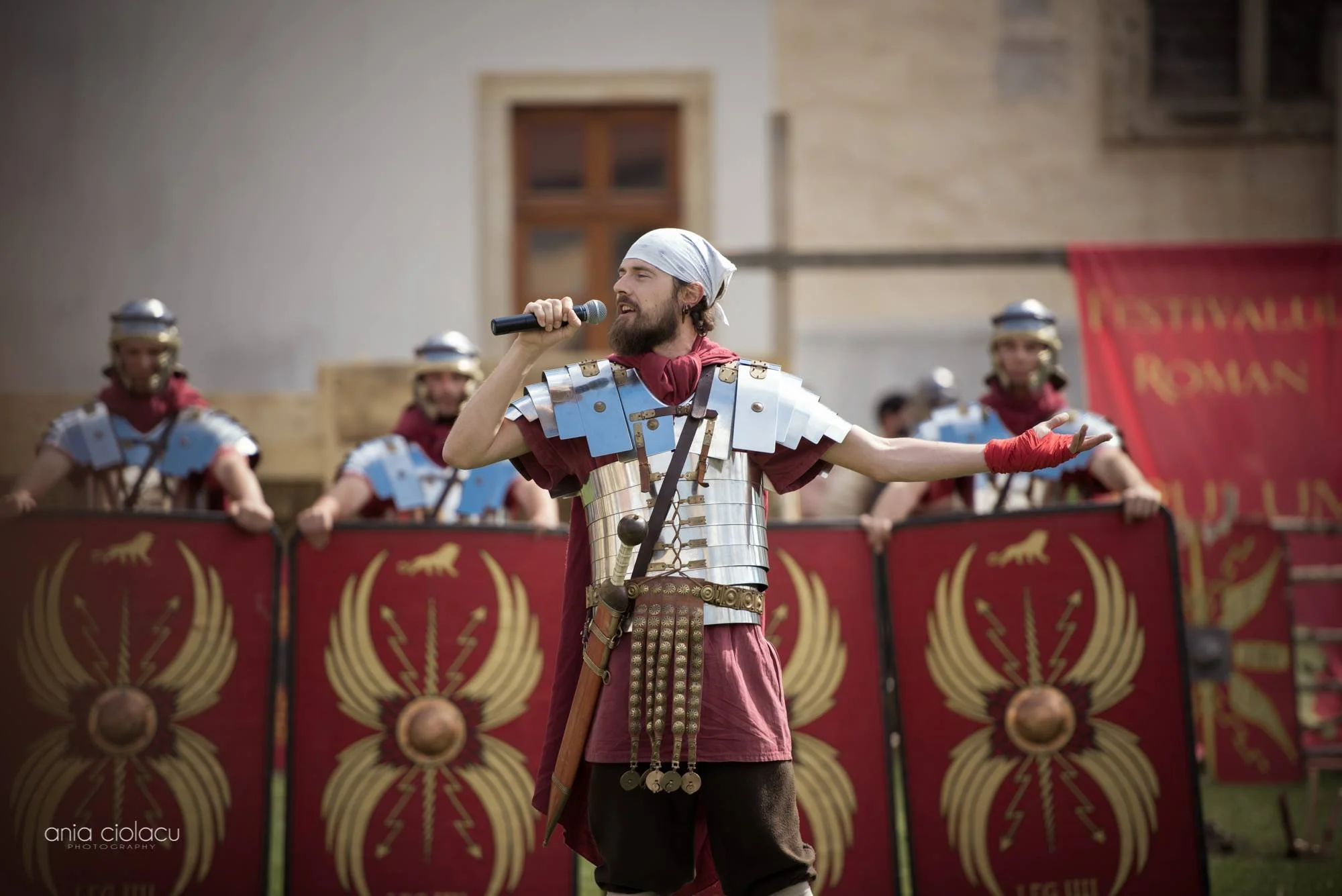 A man dressed in Roman-style armor, holding a microphone and performing in front of Roman legionary reenactors with shields and armor on a historical reenactment stage.