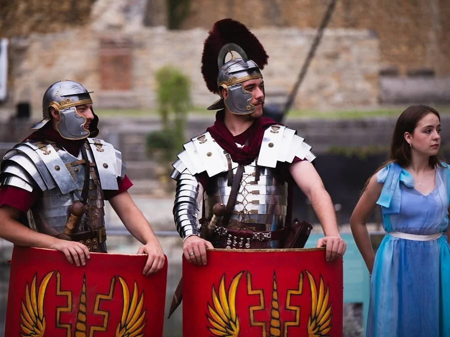 Three people dressed as Roman soldiers and a woman in a blue dress standing outdoors in front of a brick building.