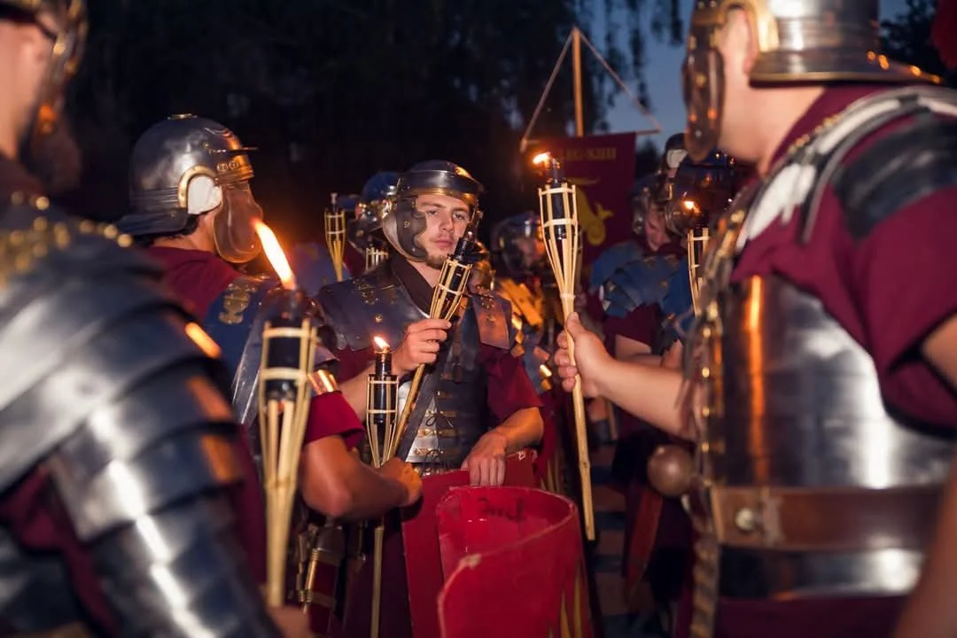 Roman soldiers dressed in historic armor, holding torches during an outdoor event at dusk.
