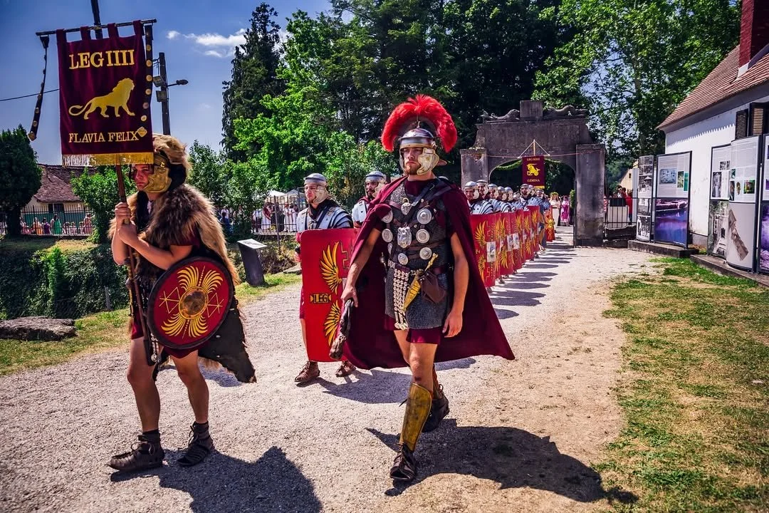 A group of people dressed as ancient Roman soldiers and generals, marching through a park-like setting with trees and a stone archway in the background, during a historical reenactment event.