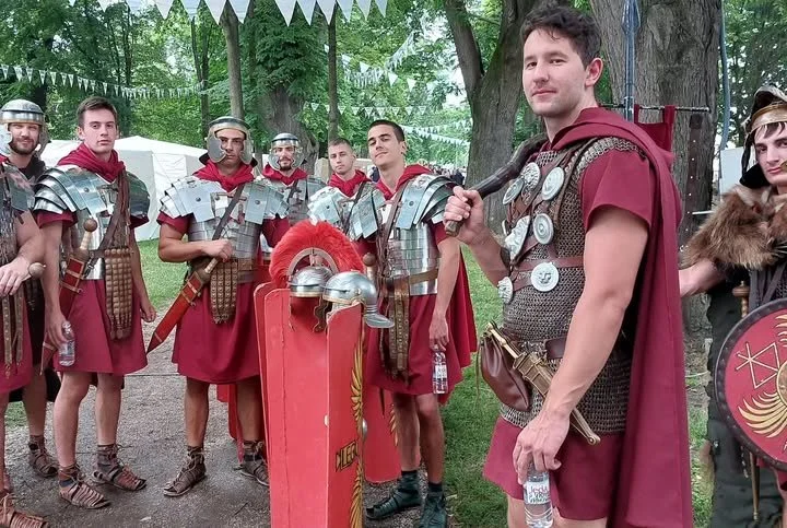 Group of young men dressed as ancient Roman soldiers, some wearing armor and holding shields, standing outdoors in a wooded area with trees and a white tent in the background.