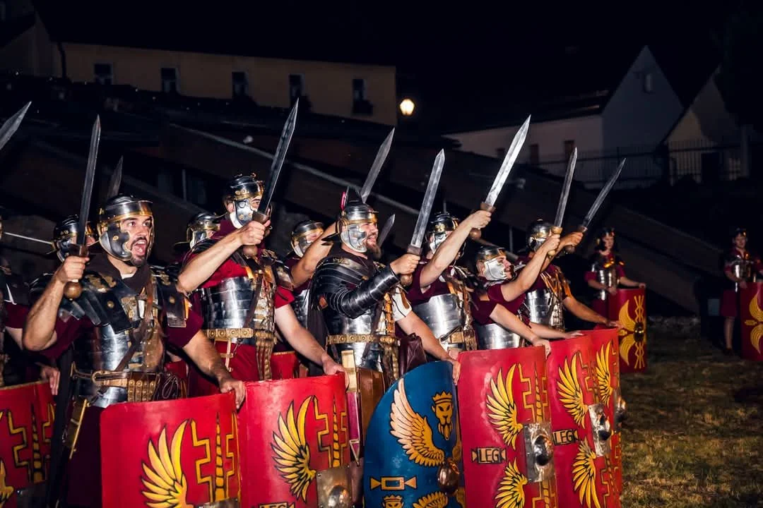 Group of people dressed as Roman soldiers holding shields and swords during a reenactment or event at night.