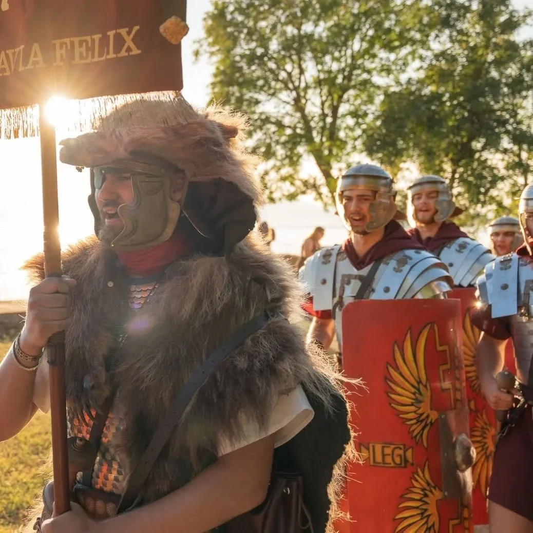 A man dressed as a Roman soldier holding a staff, wearing a traditional helmet and a fur cloak, with a group of other men dressed as Roman soldiers with shields and armor, outdoors during sunset with trees in the background.