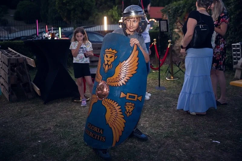 Child dressed as a Roman soldier holding a shield with a lion emblem at an outdoor event, with people and decorations in the background.