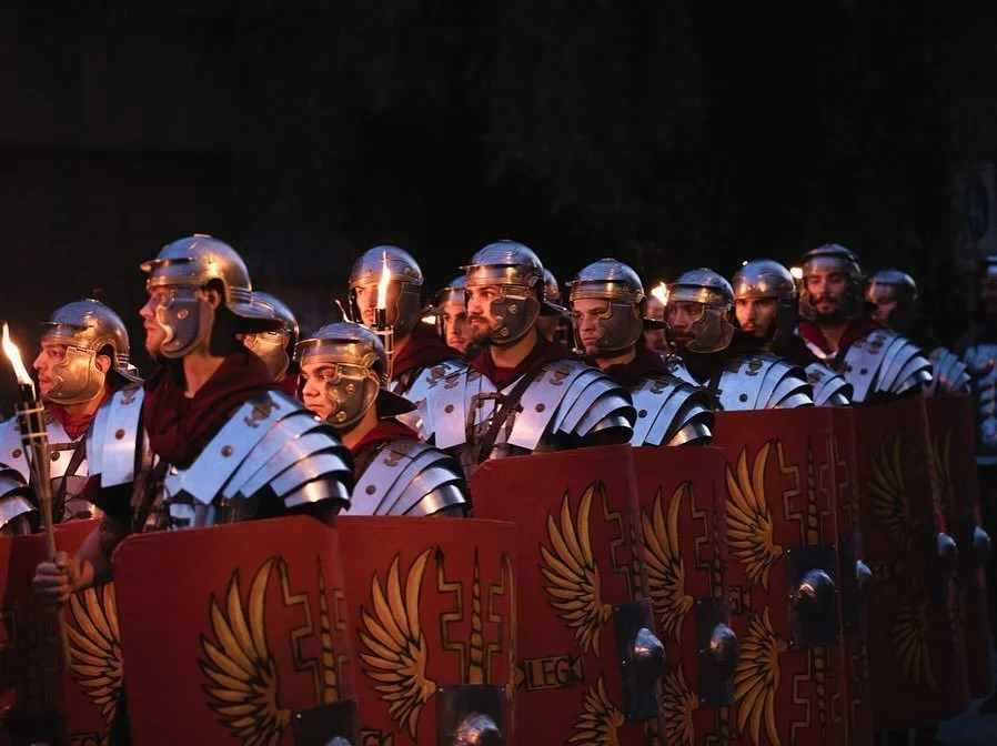 Roman soldiers in armor carrying shields with eagle emblem and torches during a nighttime event.