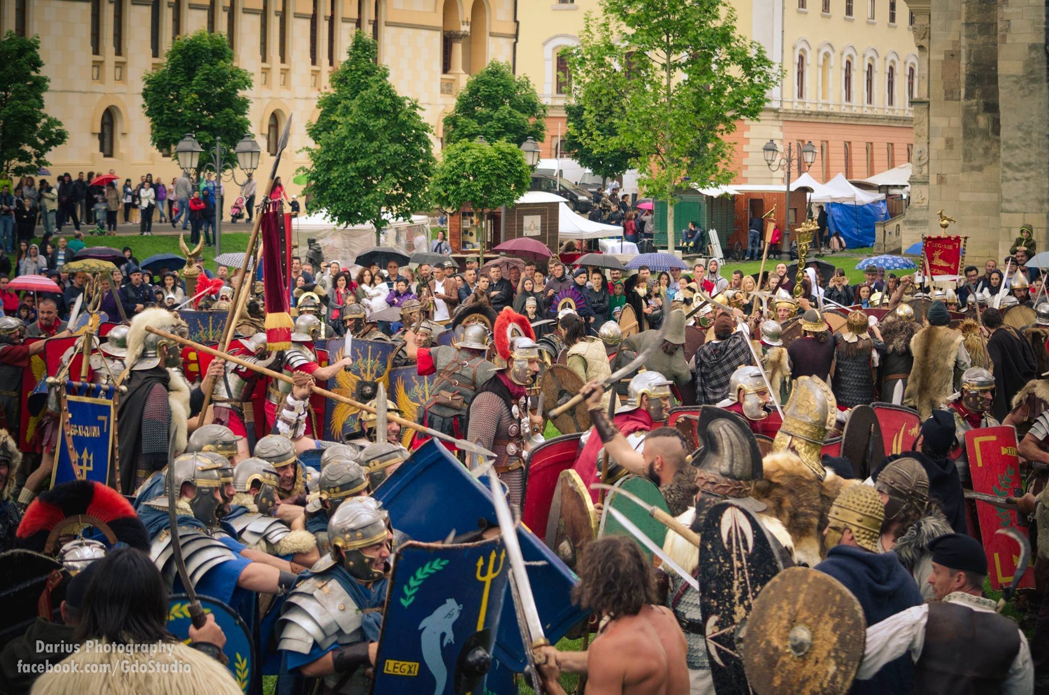 A reenactment of a historical battle with people dressed as Roman soldiers and ancient warriors, holding shields and spears in a park setting with onlookers and tents in the background.