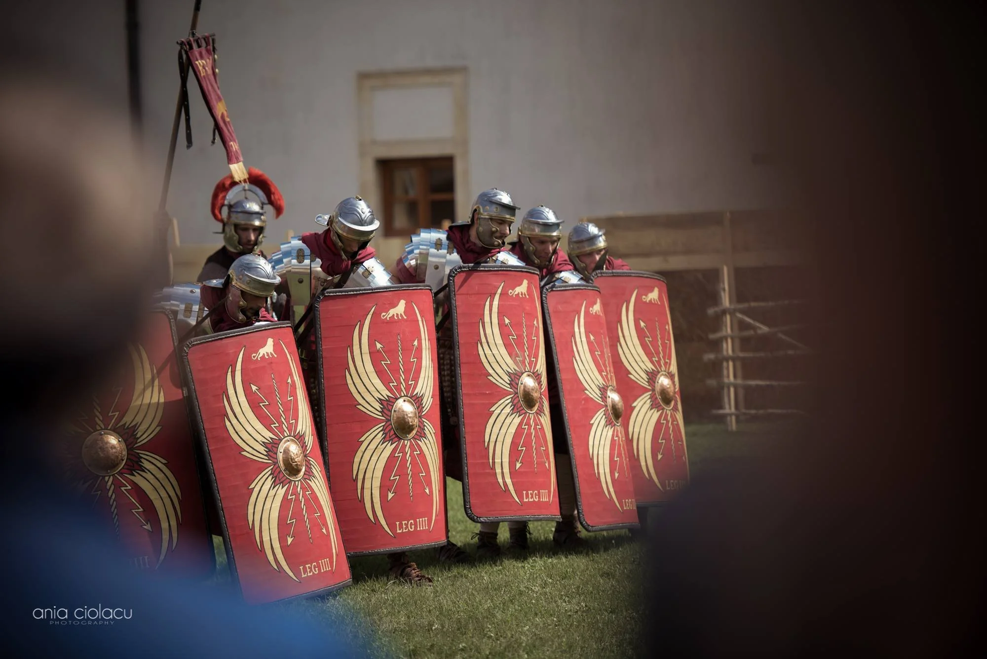 Roman soldiers standing in formation with shields featuring an eagle design, wearing armor and helmets, during a reenactment or battle scene.