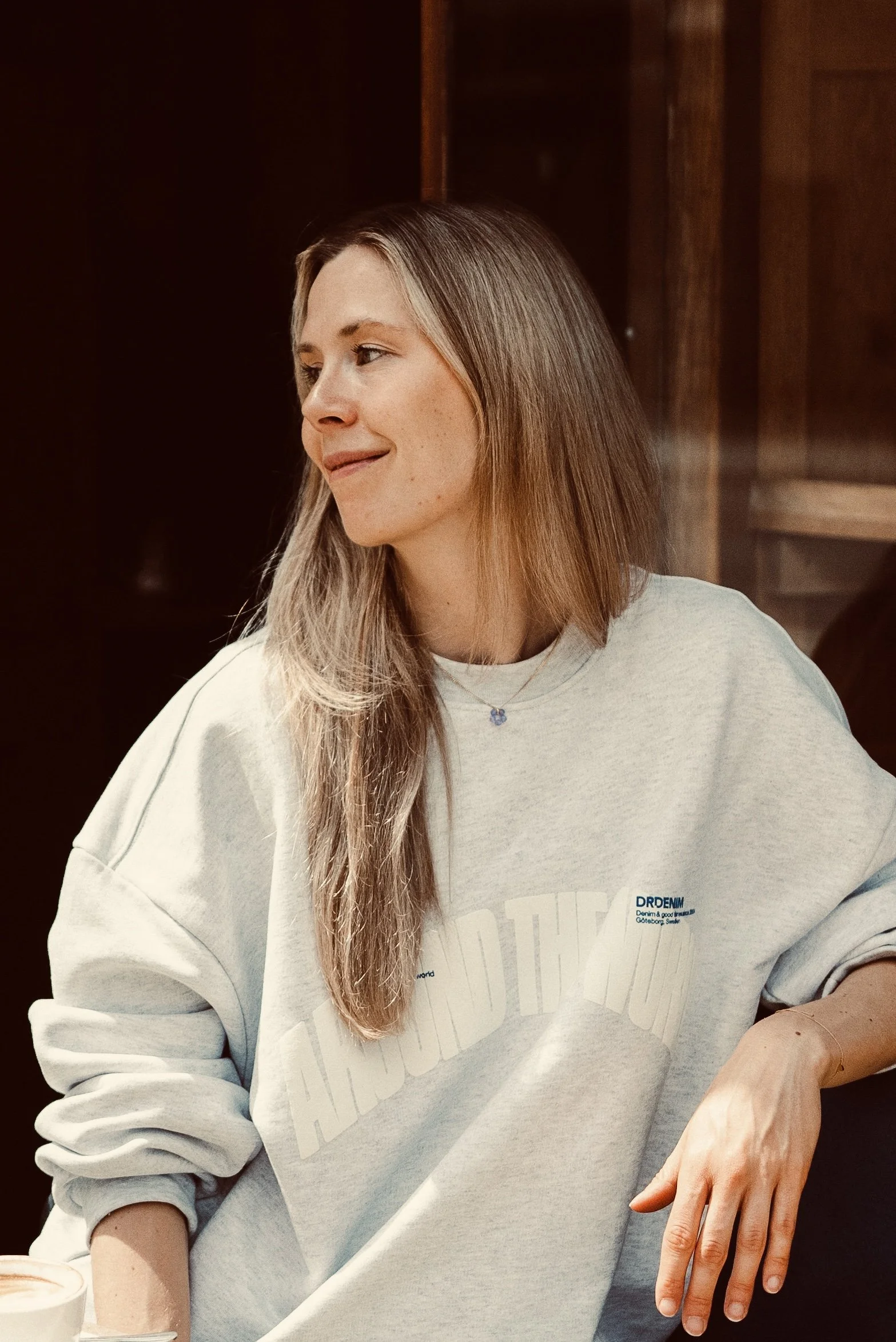 Ein Mädchen mit langem braunem Haar sitzt an einem Tisch in einem Café, trägt ein graues Sweatshirt und hält die Hand auf den Tisch. Sie hat eine kleine Kette um den Hals und lächelt leicht.