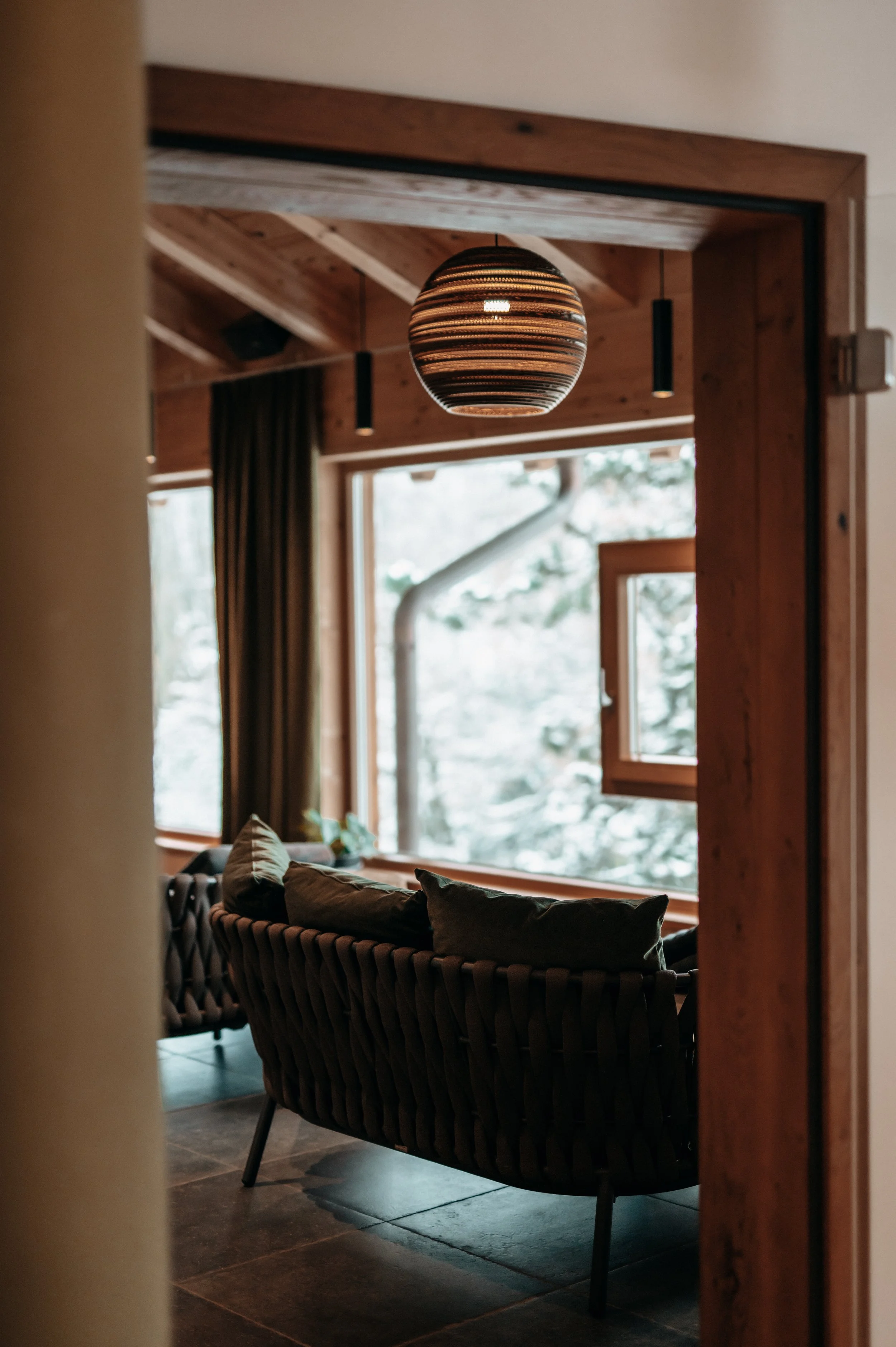 Gemütliches Wohnzimmer mit großen Fenstern, Holzdecke und einer schwarzen, gedrechselten Couch mit Kissen, im Hintergrund sind Baum und Natur sichtbar.