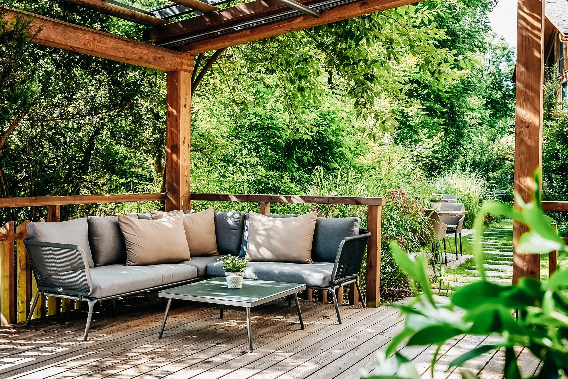 Eleganter Garten mit Holzmöbeln, bequemen Sofakissen, einem kleinen Tisch mit Topfpflanze, umgeben von üppigem Grün und einem Kiesweg im Hintergrund.