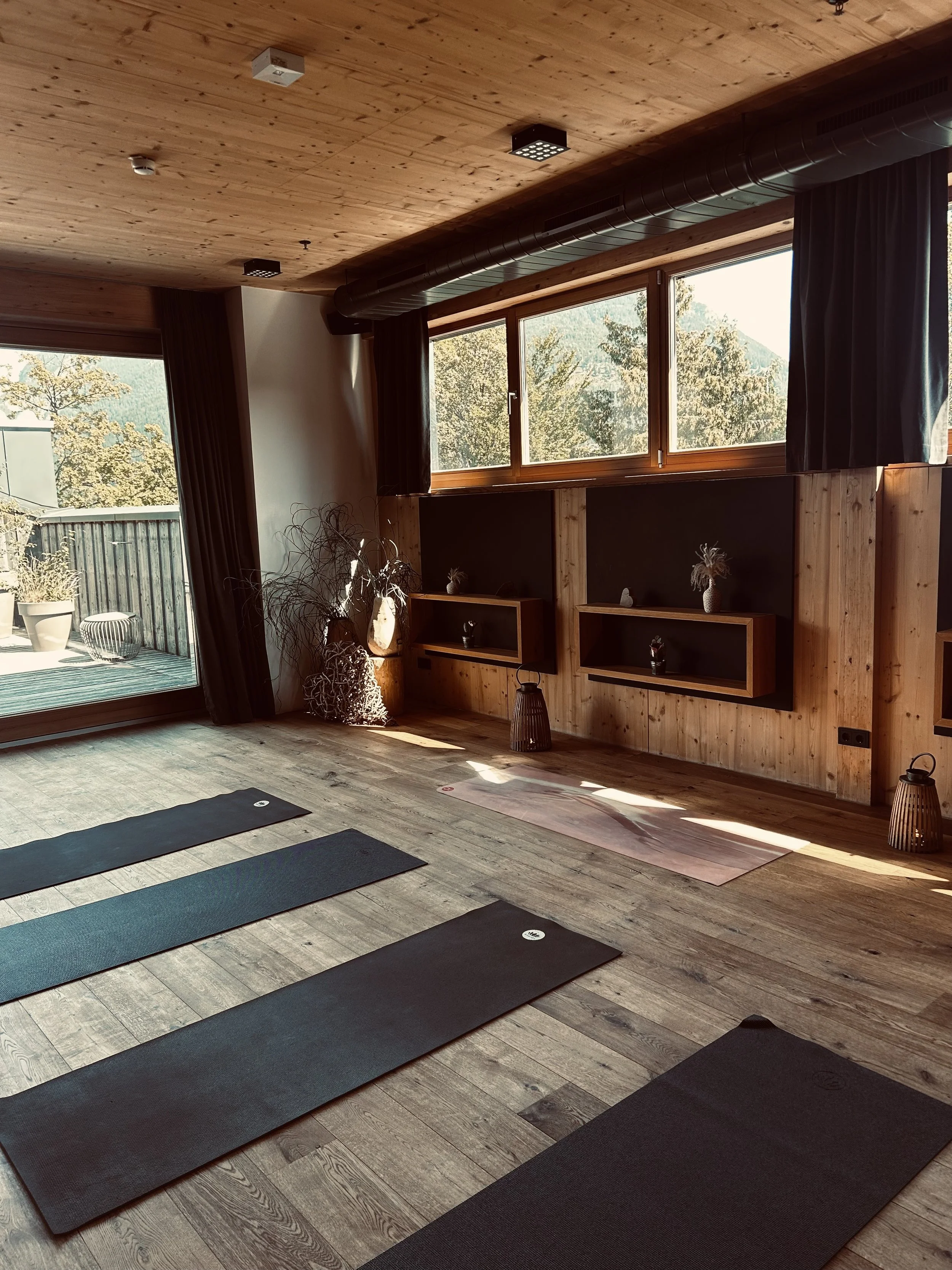 Innenraum eines Yoga-Studios mit Yoga-Matten, Holzdecke, großen Fenstern, Dekorationen und Pflanzen.