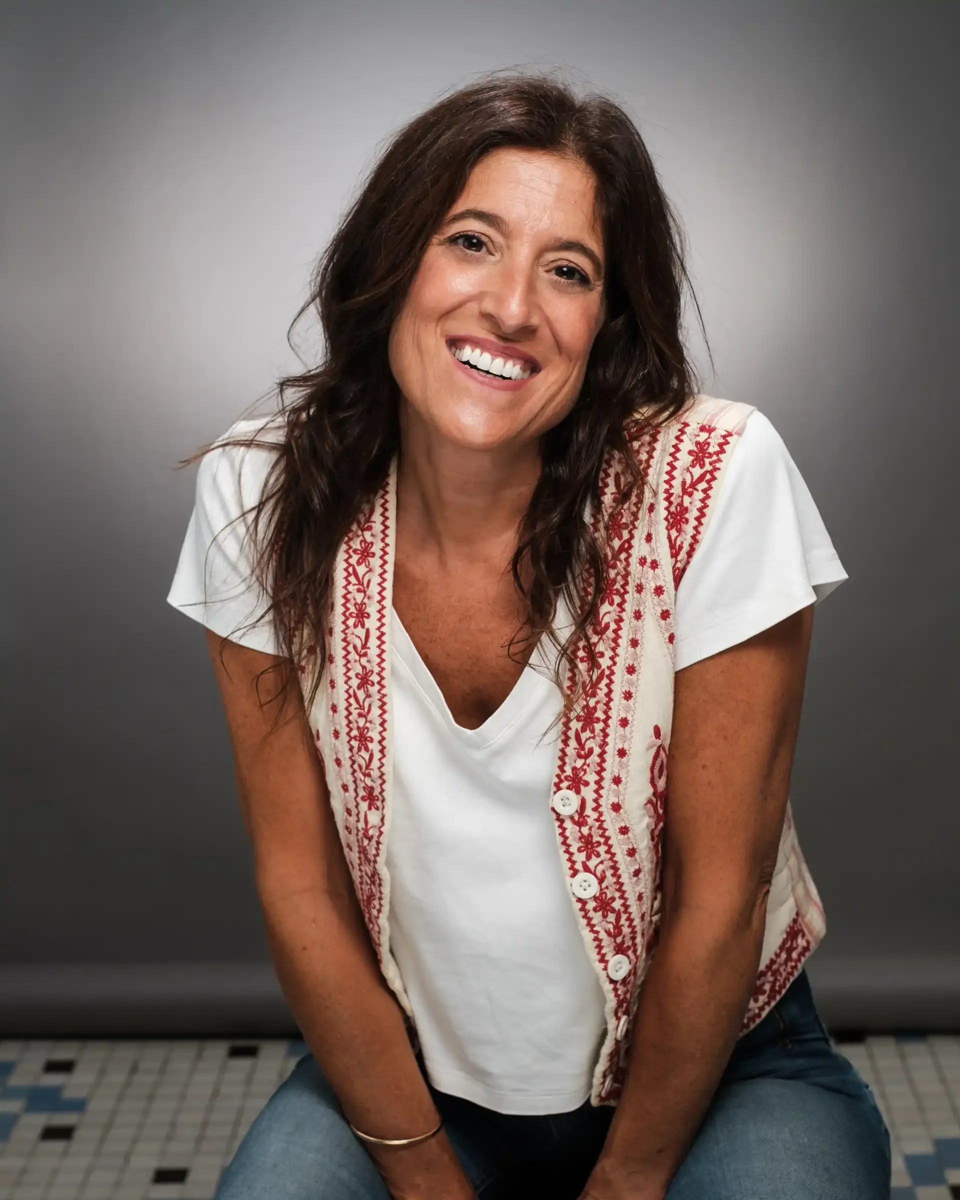Blanca Diaz, una mujer sonriendo, con cabello oscuro y rondulado, usando camisa blanca y un chaleco con bordados rojos, en un fondo gris. Usa silla de ruedas.