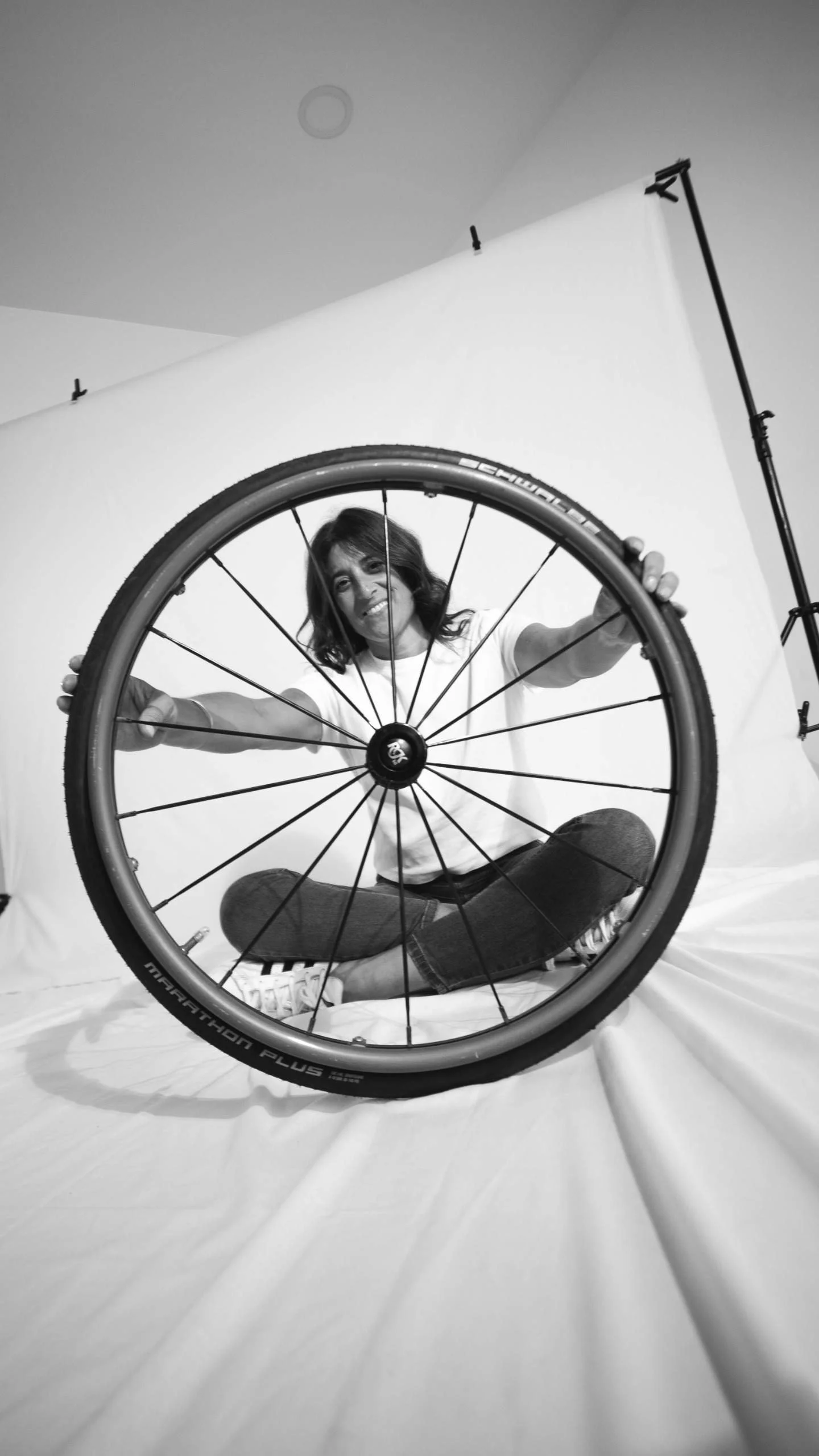 Mujer sonriente sentada en el suelo con una rueda de bicicleta frente a ella, vista desde abajo, fondo de pantalla blanco, en un estudio de fotografía.