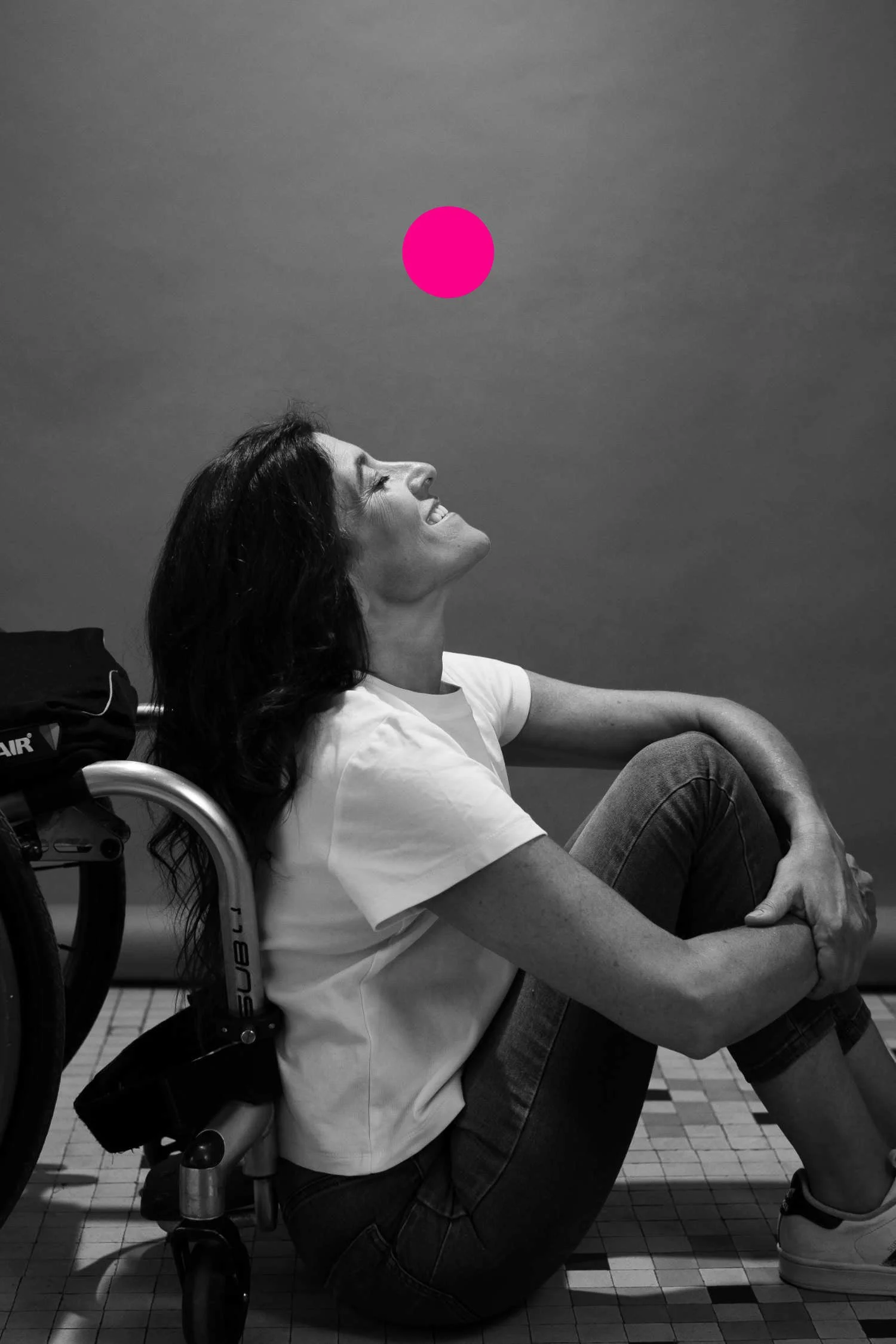 Mujer en silla de ruedas sentada en un suelo de baldosas, sonriendo, con el torso inclinado hacia atrás y las piernas dobladas hacia el pecho, mirando hacia arriba a un globo rosa flotando en el aire.